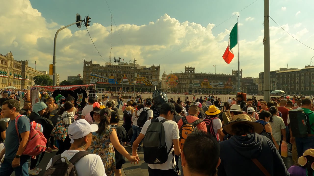 A street-level view in Zocalo Square shows a crowded foreground of pedestrians, a vendor canopy near the center, tall poles, traffic lights, and government buildings beyond the plaza.