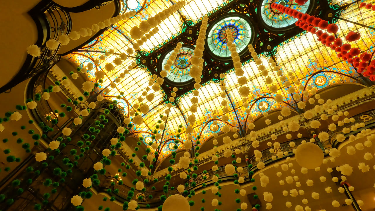Upward view inside a Mexico City building showing a glowing stained glass ceiling, circular medallions, dark ironwork, and hanging red, green, and cream decorations with no recognizable people.