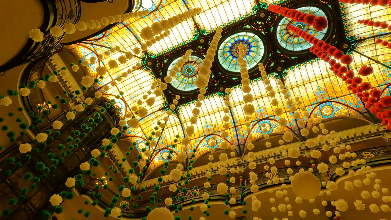 Mexico City interior ceiling frame with bright stained glass panels, blue and orange circular motifs, black structural framing, and strands of red and cream decorations crossing the view.