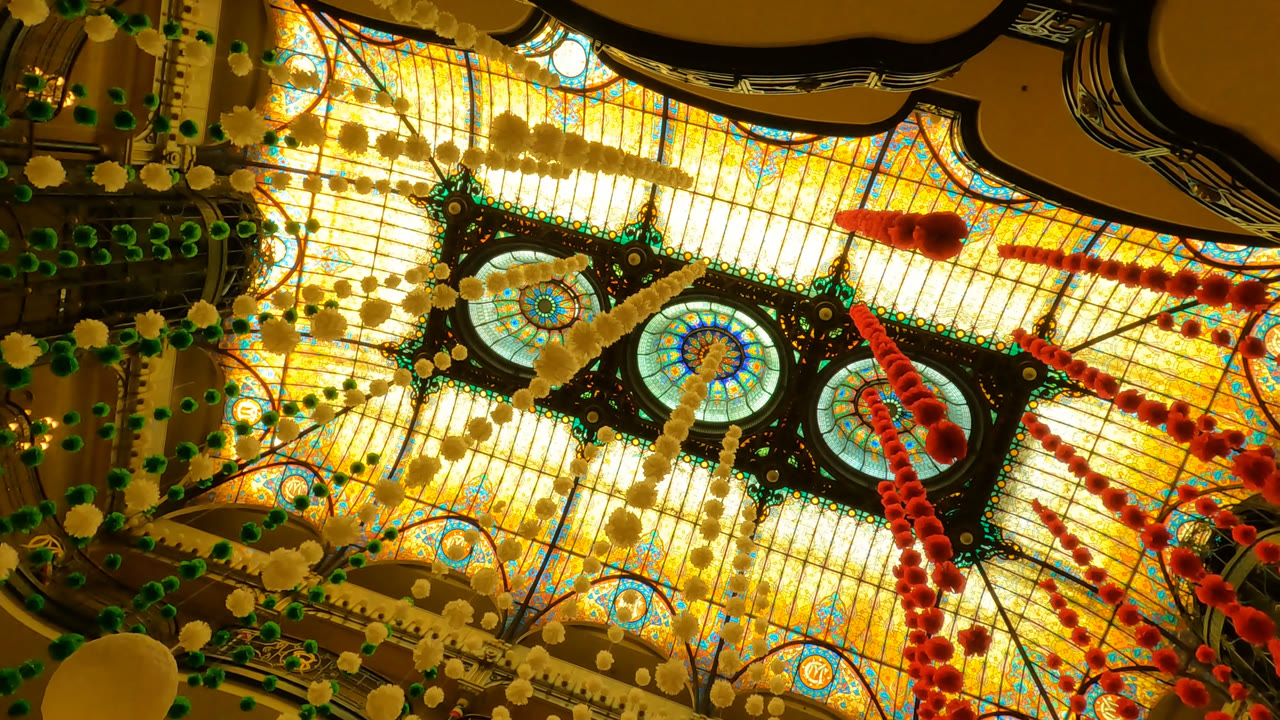 Mexico City hotel ceiling detail with two round stained glass medallions near center, warm backlighting, green and pale hanging decorations, and no visible people.