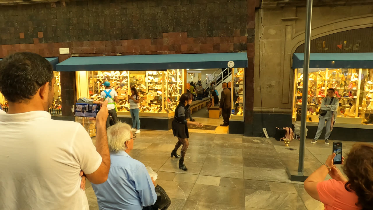 In Mexico City, spectators stand and sit in the foreground while a black-clad street performer faces a brightly lit storefront display across the sidewalk.