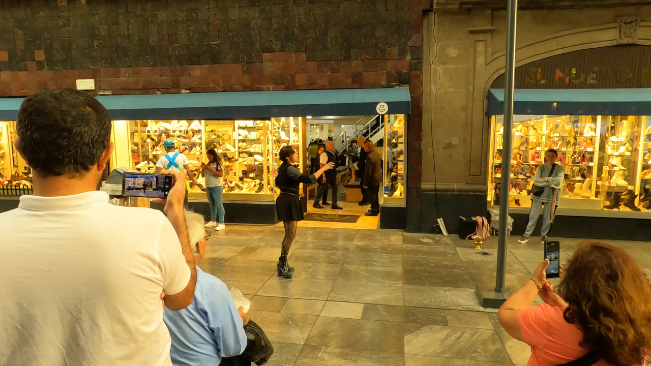 In Mexico City, a man in a white shirt holds up a phone to record as the performer stands in the sidewalk space near the illuminated shop window.