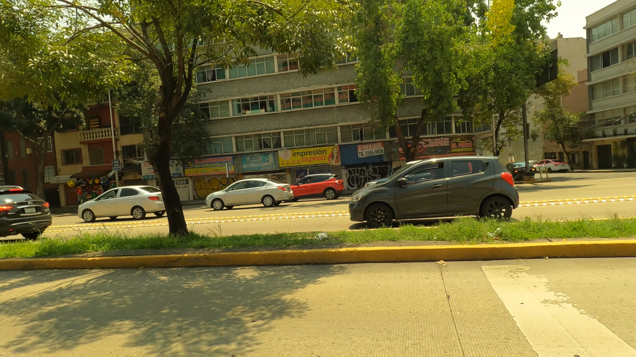 Opening frame of Mexico City traffic with a gray hatchback, smaller cars, tree shade, storefront signs, multi-story buildings, and no visible people.