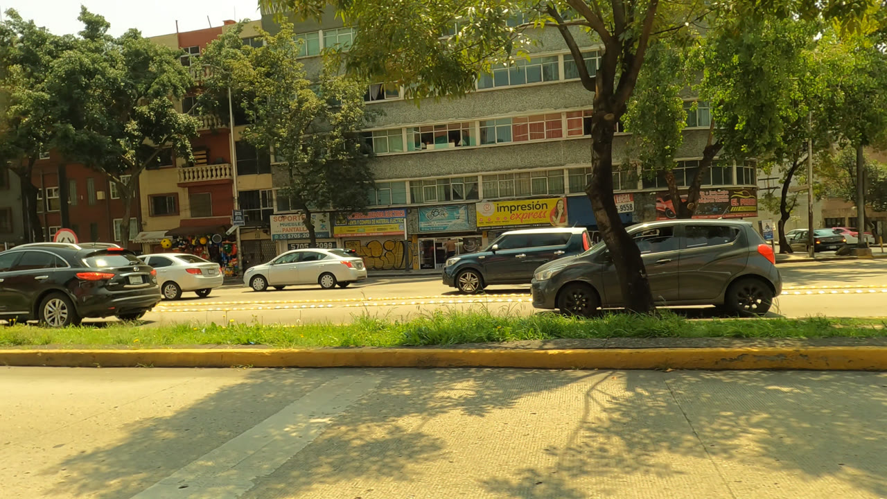 Mexico City street view with black and gray vehicles crossing the frame, a grassy median, storefront signs, apartment-like windows, and no visible people.