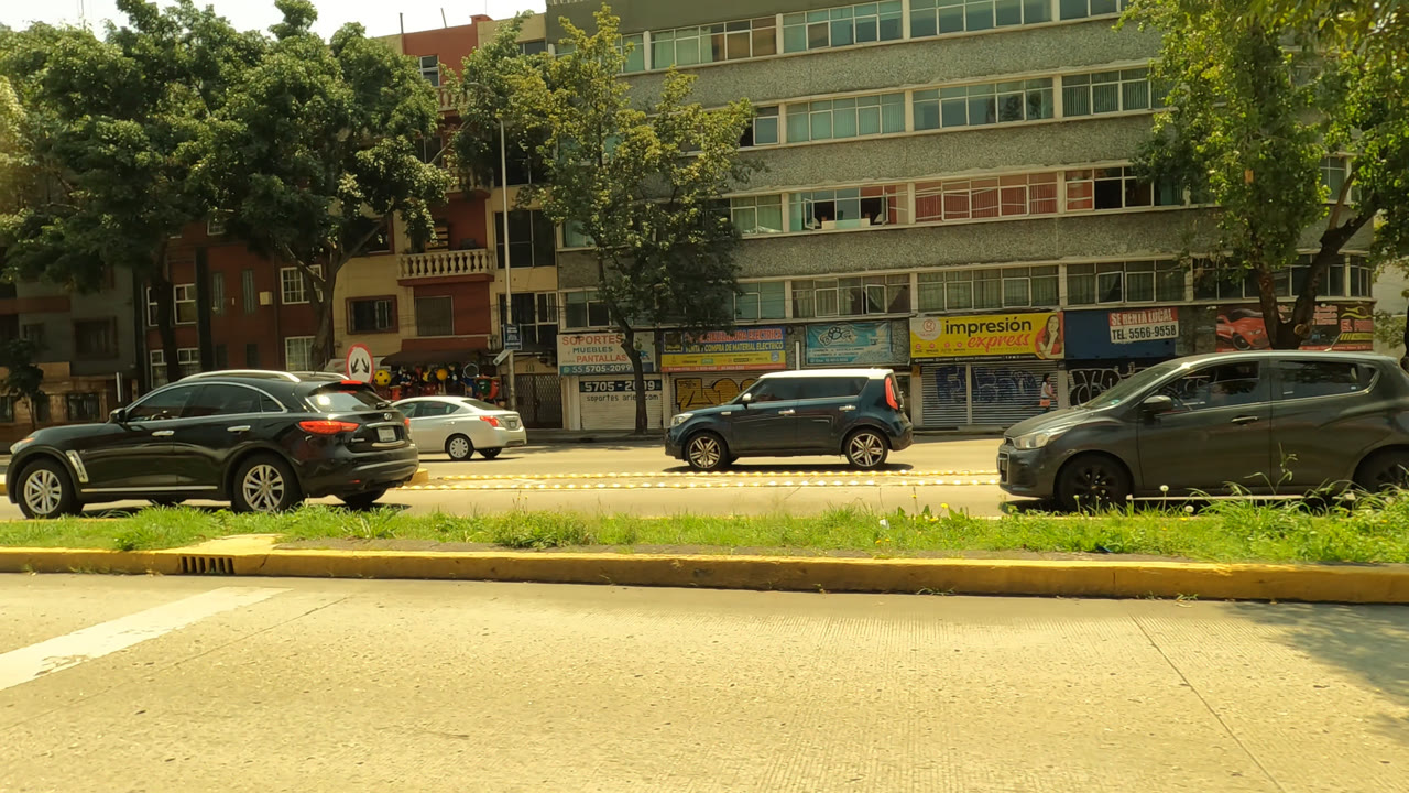Traffic continues along the Mexico City roadway with a dark sedan, white compact car, gray hatchback, leafy trees, storefront awnings, and no visible people.