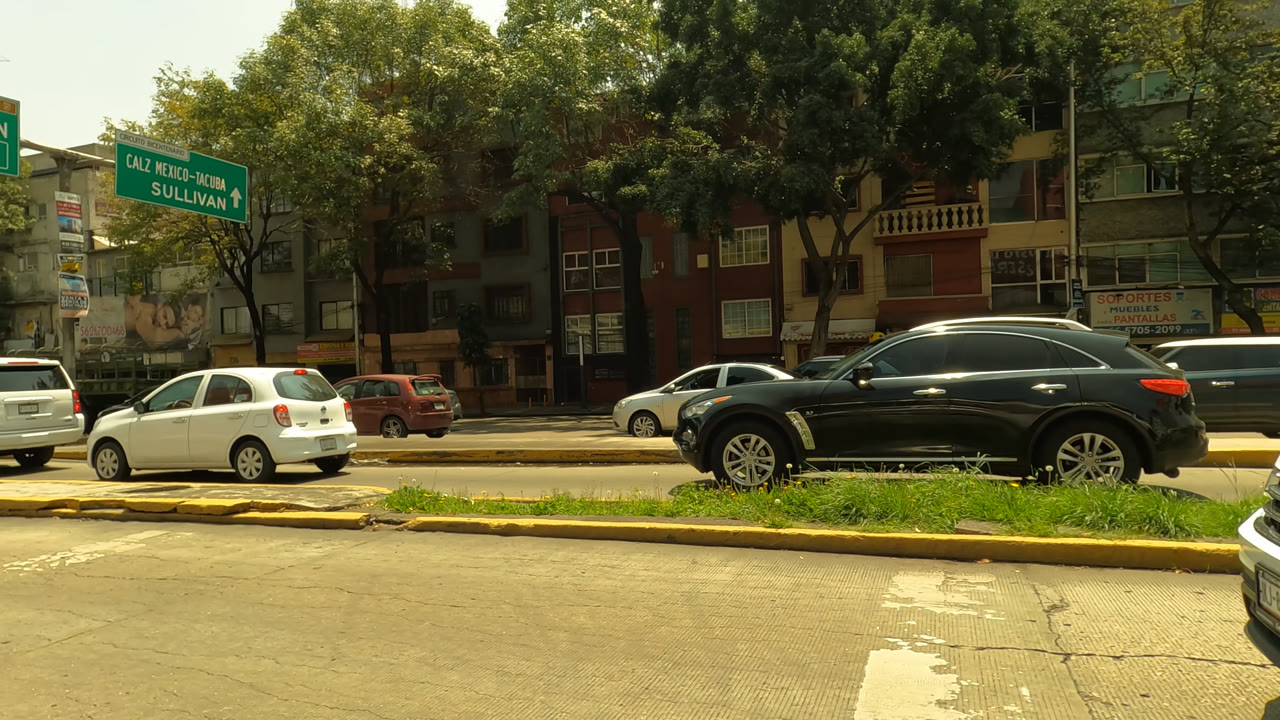 Opening frame of a Mexico City road with a black SUV, white cars, yellow curb barriers, leafy trees, apartment buildings, green street signs, and no visible people.