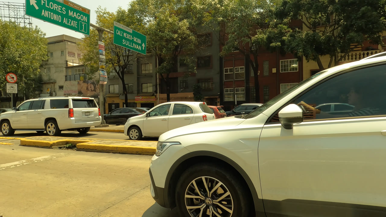 The white SUV fills the right side of the Mexico City frame, with its front wheel and side visible against a background of parked cars, trees, buildings, and green street signs, with no visible people.