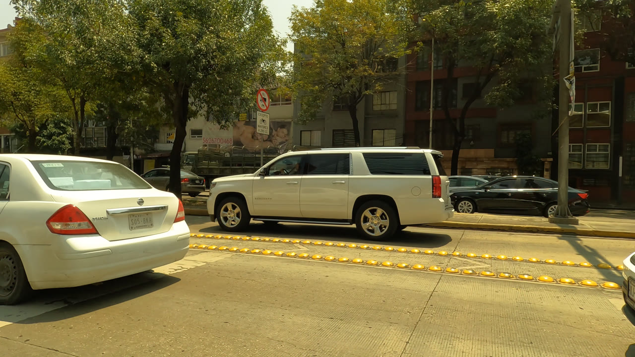Street-level Mexico City frame showing a white sedan in the foreground, a white SUV across the median, trees, apartment buildings, graffiti, and no visible people.