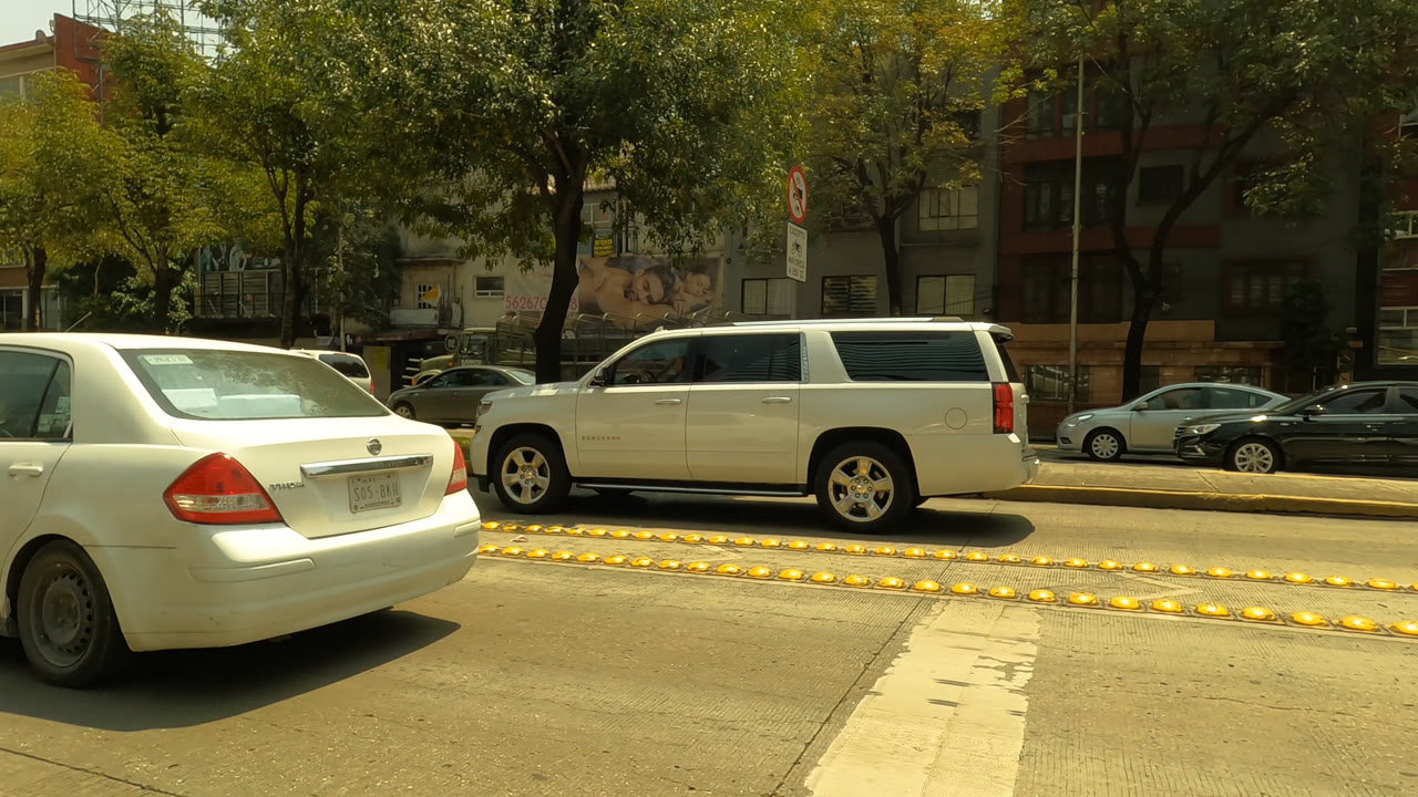 Mexico City traffic view with the rear of a white sedan near the camera, a white SUV in the opposite lane, parked cars, trees, wall art, and no visible people.