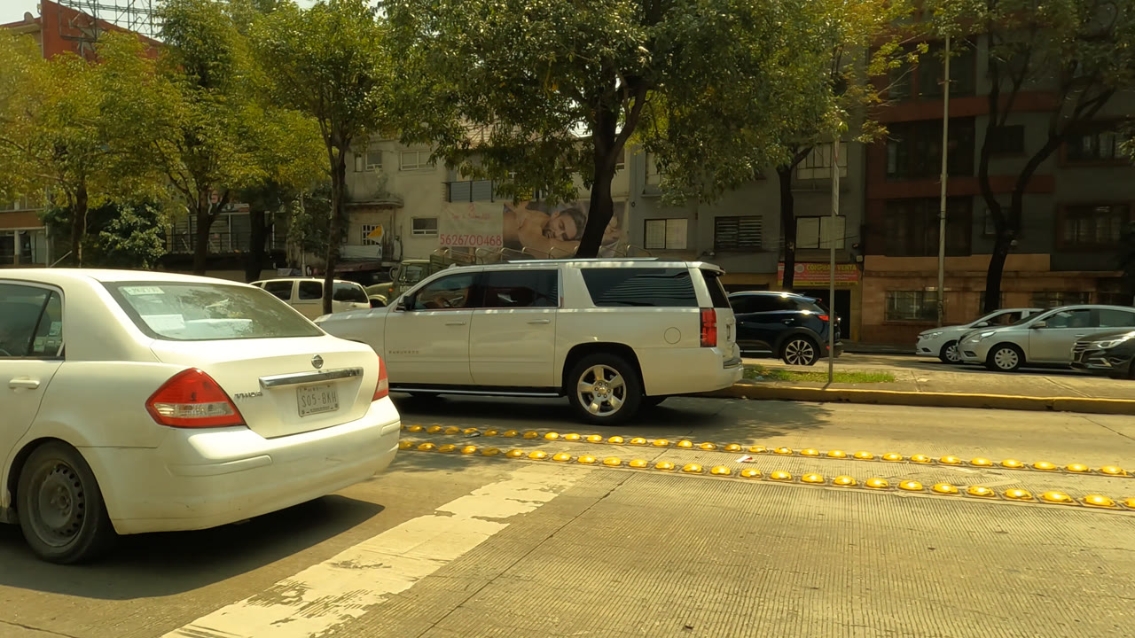 Mexico City street frame with a white sedan angled across the foreground, a white SUV beyond the divider, shaded trees, parked cars, building facades, and no visible people.