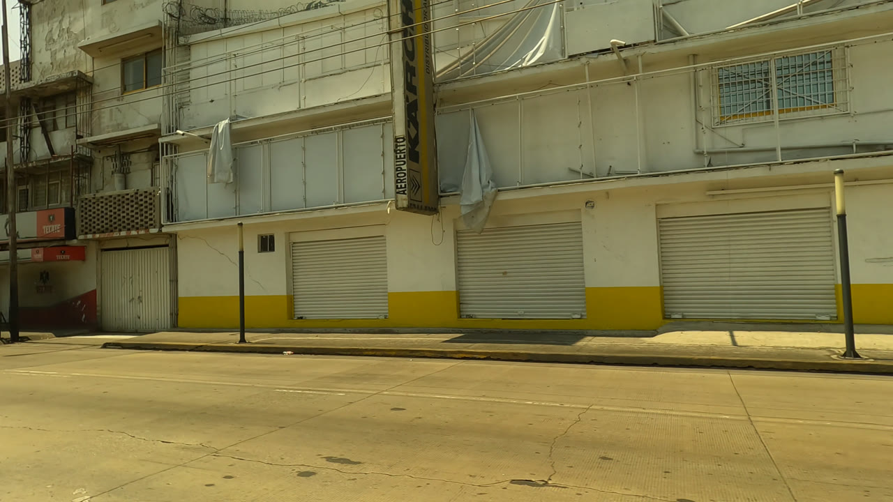 Sunlit city street with closed storefronts, apartment balconies, trees, and overhead wires.