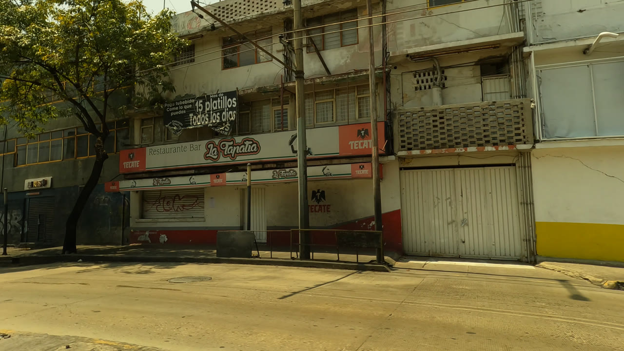 Mexico City street frame with shuttered storefronts, graffiti on a blue wall, trees, apartment levels, and an orange medical lab advertisement on a building.