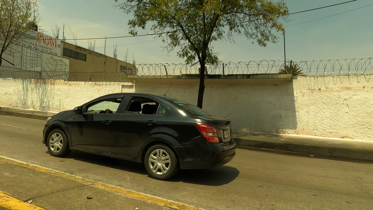 The black sedan continues along the Mexico City road beside a long white wall topped with barbed wire, with trees, wires, and a low industrial building behind it.
