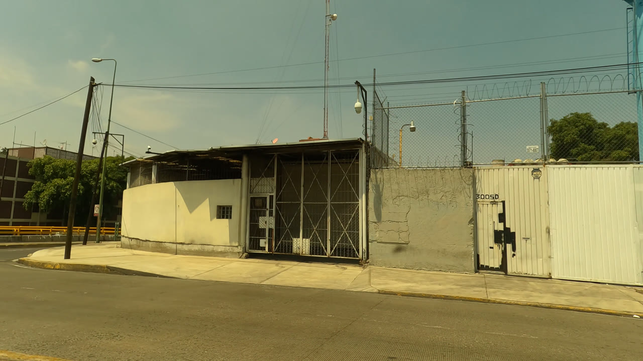 Mexico City street frame with a white metal gate, small guard structure, utility poles, yellow roadside barrier, and no visible people.