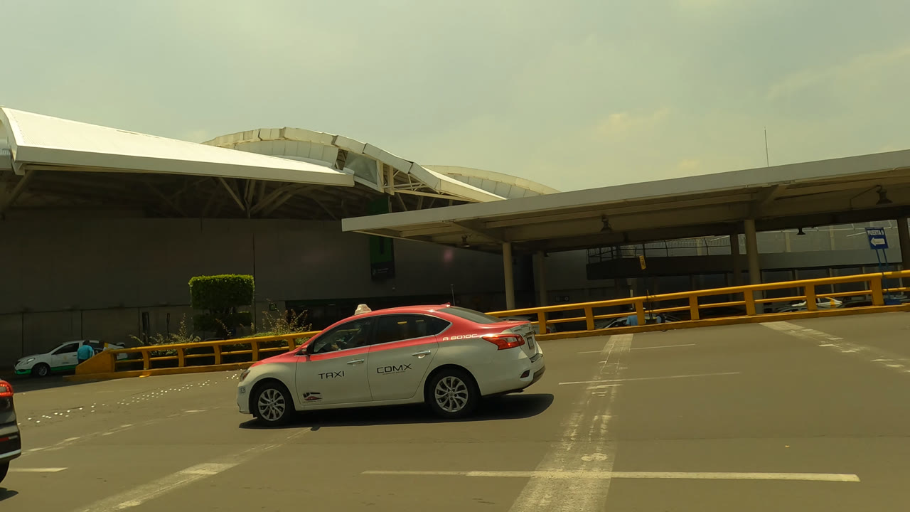 The taxi is centered in the lane beside a Mexico City airport terminal wall, with yellow guardrails, parked vehicles, and no clearly visible people in this frame.