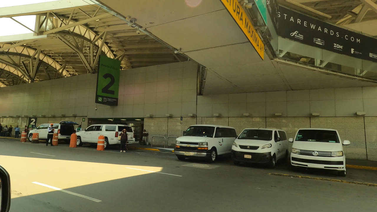 Mexico City airport curbside pickup area with white vans, SUVs, orange traffic barrels, a green number 2 sign, shaded terminal wall, and small people along the sidewalk.