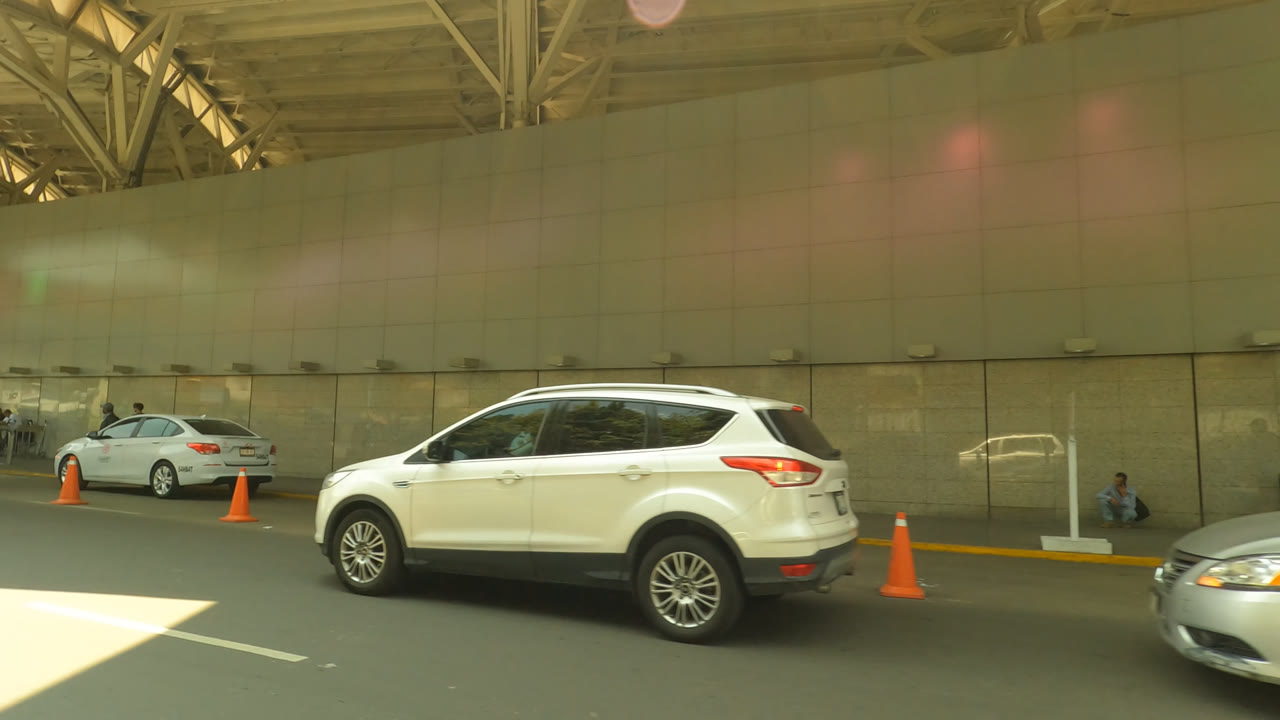 Mexico City airport curb with a white SUV centered near orange cones, a hanging green number 3 terminal sign, beige facade panels, and small distant travelers.