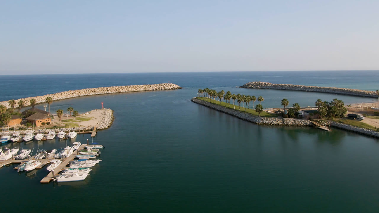 Aerial frame over Puerto Los Cabos marina with docked white boats at lower left, dark harbor water, pale rock breakwaters, palm trees, open ocean, and no visible people.