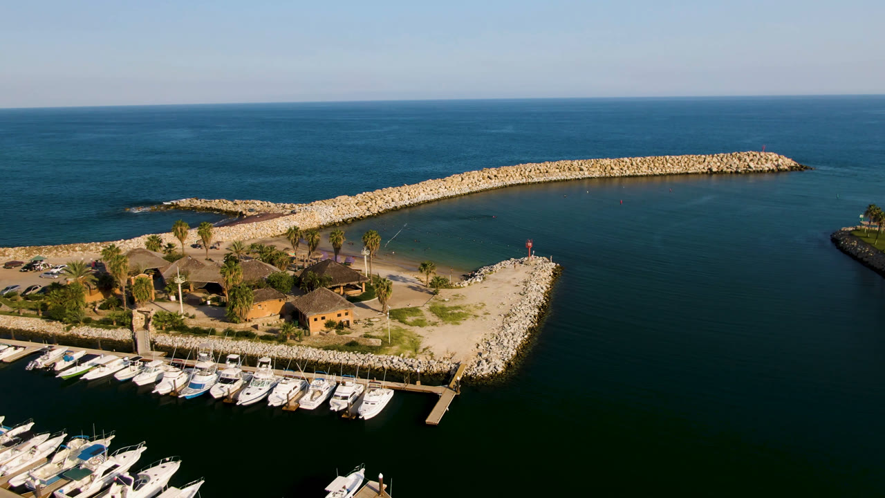 Opening aerial frame over Puerto Los Cabos shows docked boats at the lower edge, a small palm-lined island, a curved rock breakwater, open ocean, and no visible people.