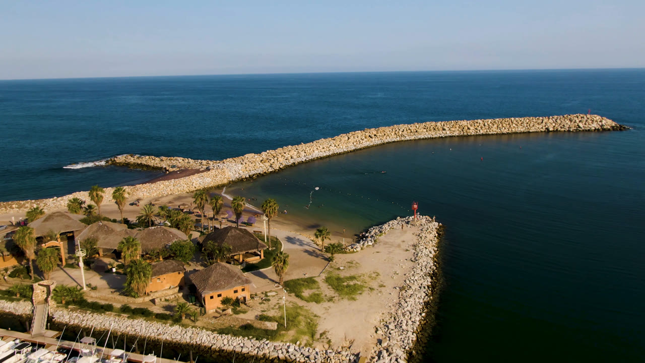 Opening aerial frame over Puerto Los Cabos with thatched-roof buildings and palms at lower left, a sandy point with a red beacon, curved rock breakwater, blue-green water, and no visible people.
