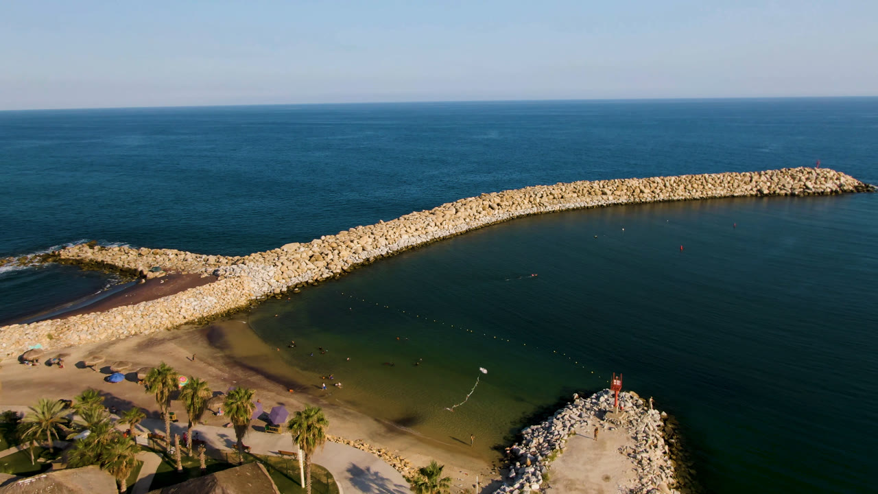 Aerial view of a curved rock breakwater, beach, palms, and open ocean.