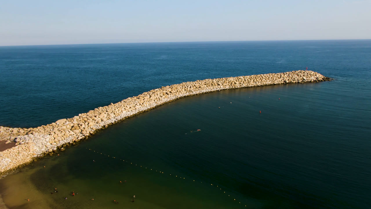 Aerial frame from Puerto Los Cabos showing a long pale stone breakwater curving across blue-green water with open ocean and no visible people.
