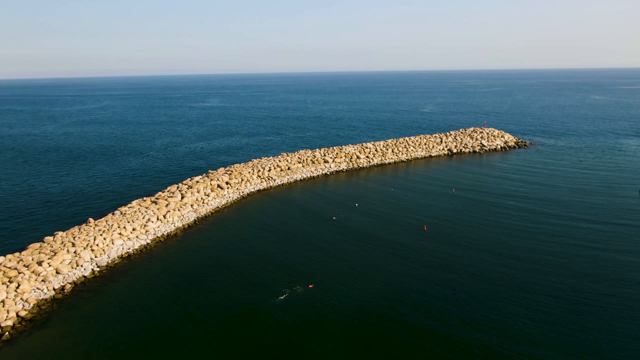 Aerial frame from Puerto Los Cabos showing a curved pale rock breakwater crossing dark green ocean water with open blue sea beyond and no visible people.