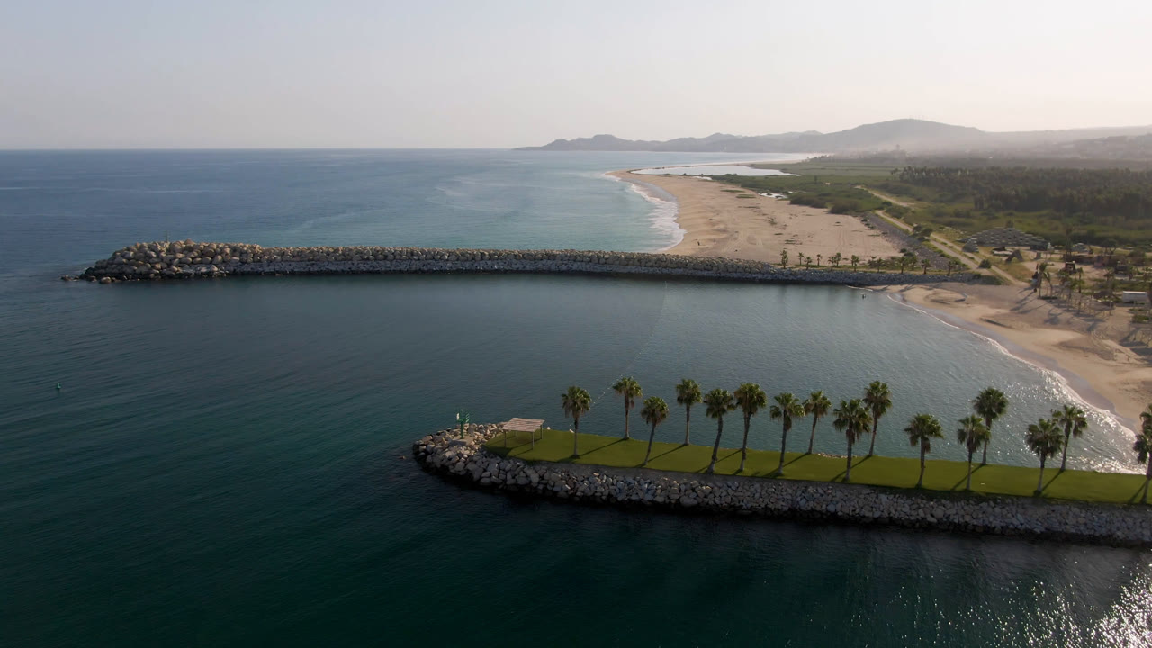 Aerial frame over Puerto Los Cabos showing dark blue water, a long rock pier, pale beach, a foreground rocky point with palms, distant hills, and no visible people.