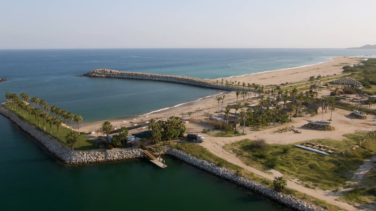 Aerial frame over Puerto Los Cabos showing blue-green water around rock breakwaters, a sandy beach beyond the inlet, palm rows, small dock structures, and no visible people.