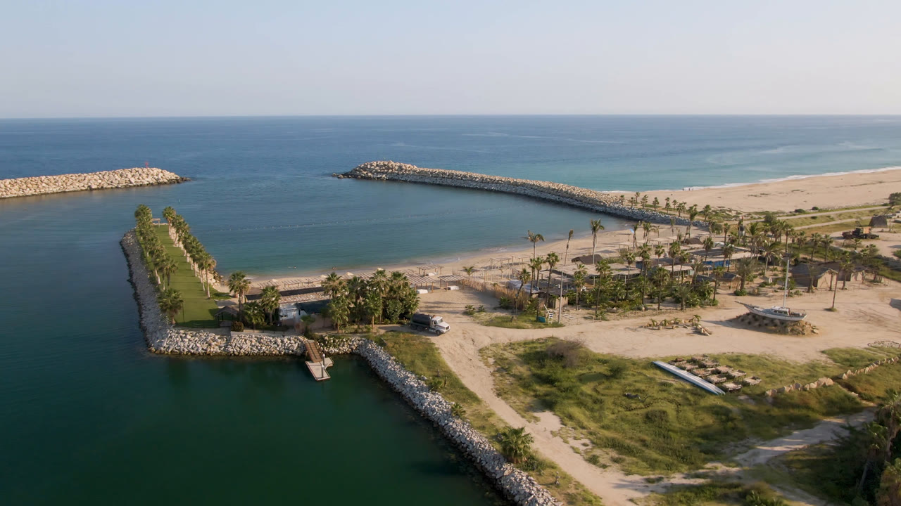 Opening aerial frame over Puerto Los Cabos with dark turquoise water in the foreground, rock breakwaters framing a sandy beach, palms inland, and no visible people.