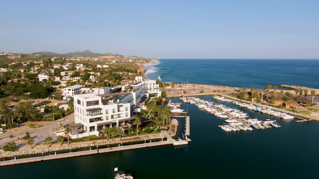 Aerial view of a marina with docked boats, a white waterfront building, ocean, and hillside homes.