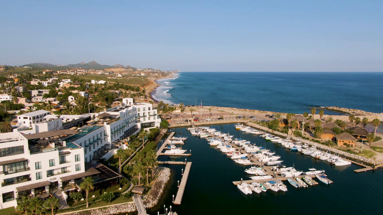 Opening aerial frame of Puerto Los Cabos with white boats in a marina, waterfront buildings at left, surf along the coastline, blue ocean to the right, and no visible people.