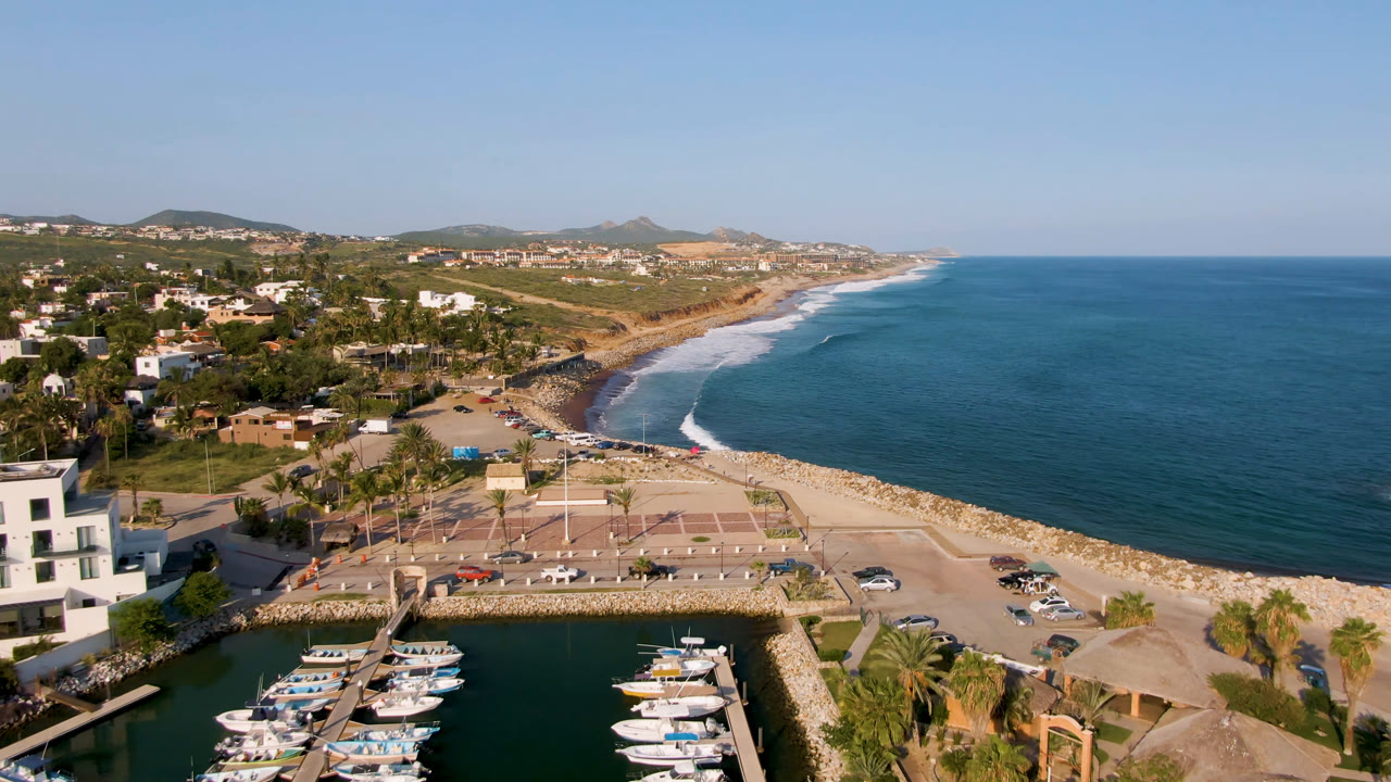 High aerial view over Puerto Los Cabos with docked boats in the marina foreground, a paved waterfront plaza, surf along the beach, low hills in the distance, and no visible people.