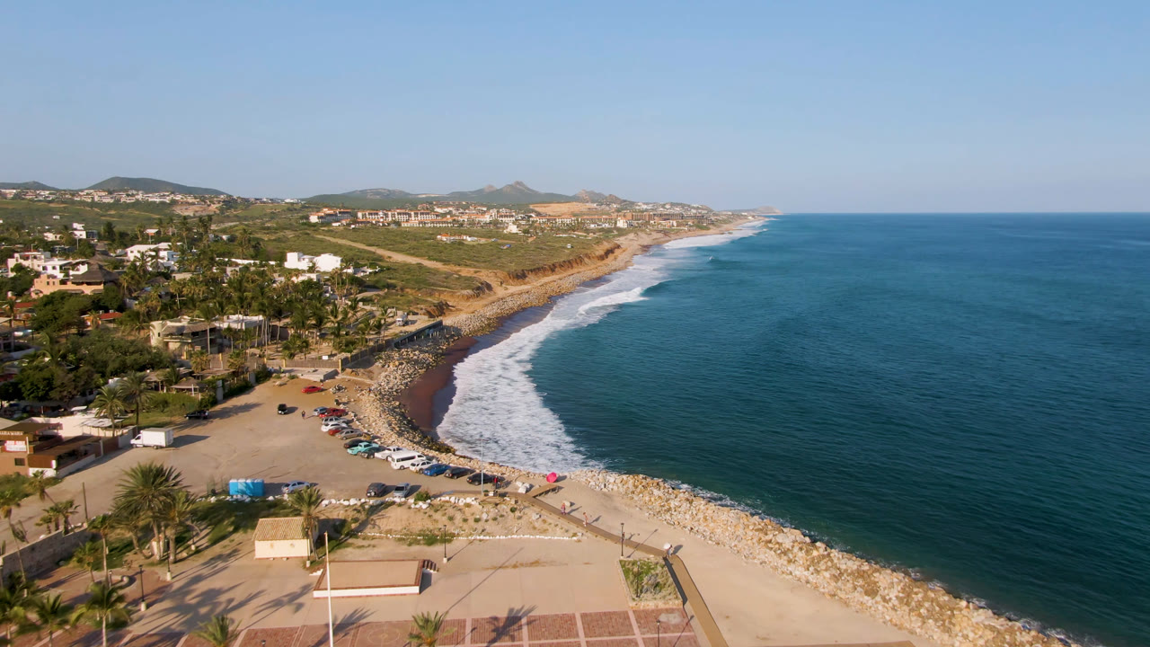 Aerial view of curved beach, surf, parked cars, palms, homes, hills, and blue ocean.