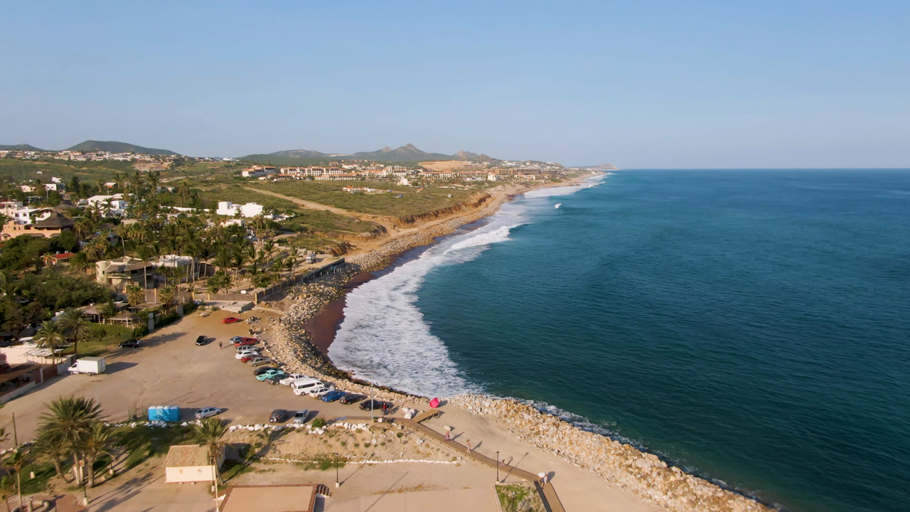 One second into the Puerto Los Cabos clip, the aerial view holds on white surf wrapping the rocky shoreline, with beach parking, palm trees, homes, hills, and no visible people.