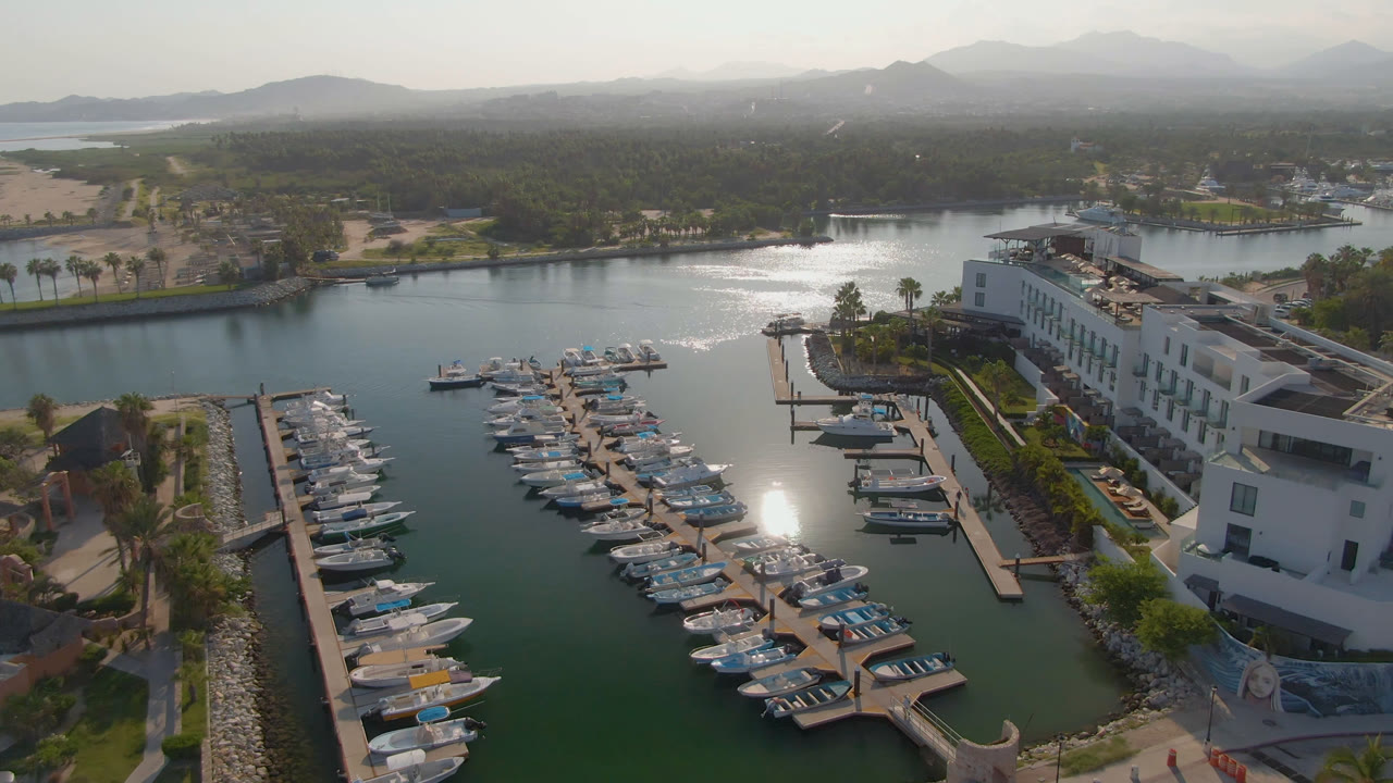 Opening aerial frame over Puerto Los Cabos shows docked boats in green-blue marina water, a white waterfront hotel, hazy mountains, and no visible people.