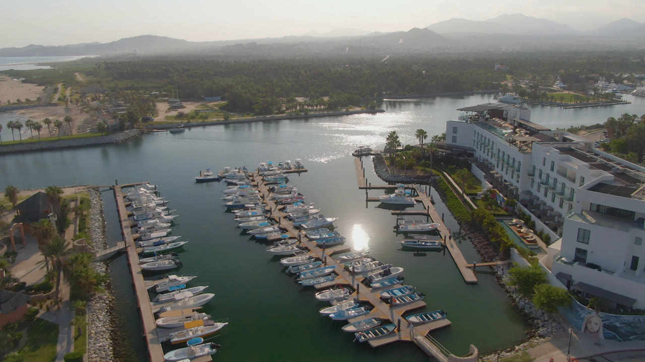 Wide aerial marina scene with boats, docks, hotel buildings, palms, and mountain backdrop.
