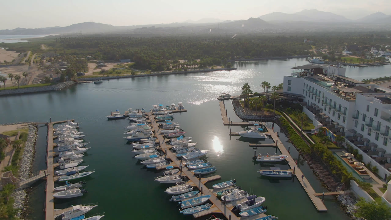Aerial frame from Puerto Los Cabos showing rows of boats at marina docks, a white resort building along the water, hazy hills beyond, bright water glare, and no visible people.