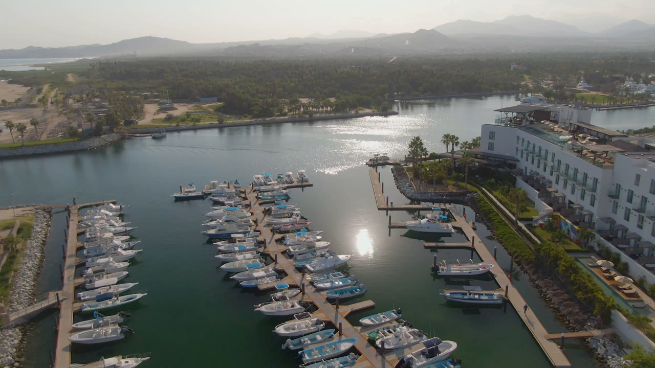 Puerto Los Cabos marina view with clustered white boats in the foreground, the resort building on the right, calm water reflecting the sun, distant hills, and no visible people.