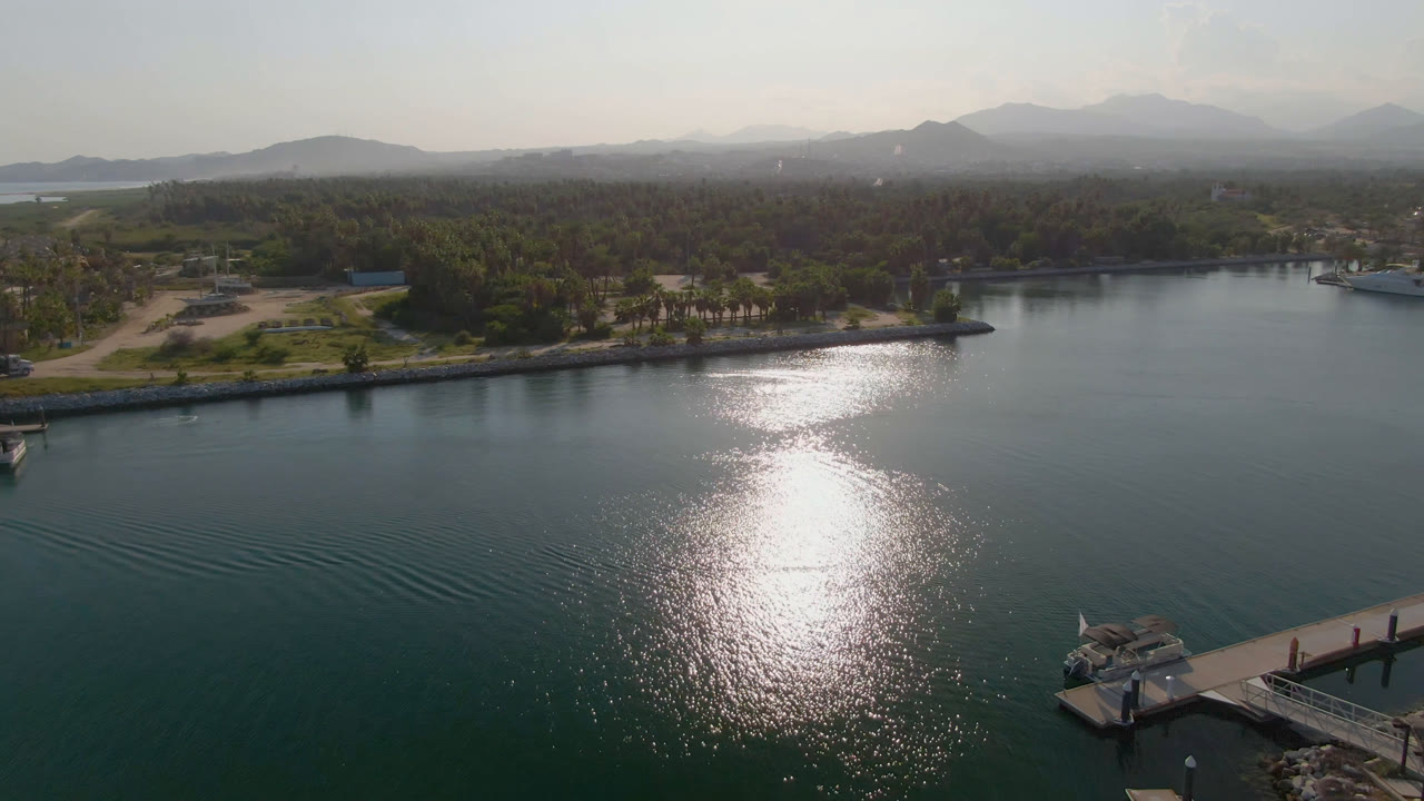 Aerial frame over Puerto Los Cabos water with bright sun reflection in the foreground, a dock at the lower right edge, palm-lined shore beyond, distant mountains, and no visible people.