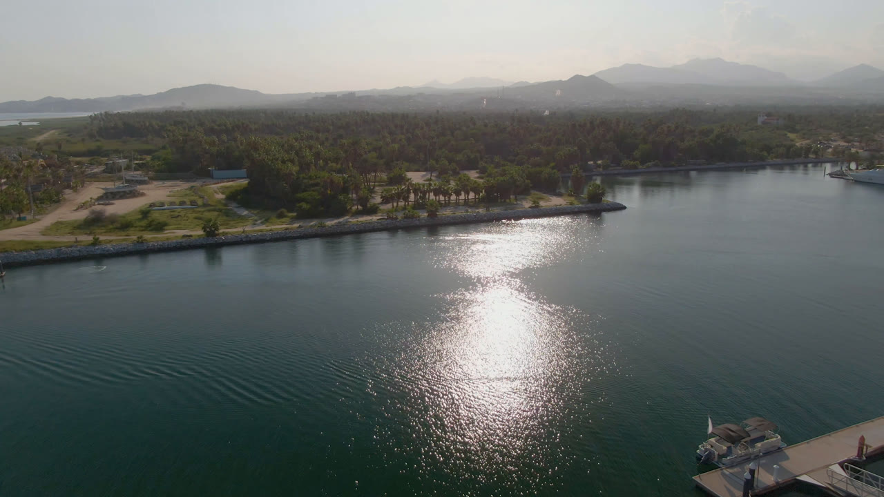 Aerial frame over Puerto Los Cabos water where the sun reflection sits near the center, with a low shoreline, palms, small structures, hazy mountains, and no visible people.