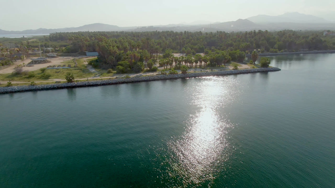 Aerial shoreline view with calm water, bright reflection, palms, and mountains in haze.