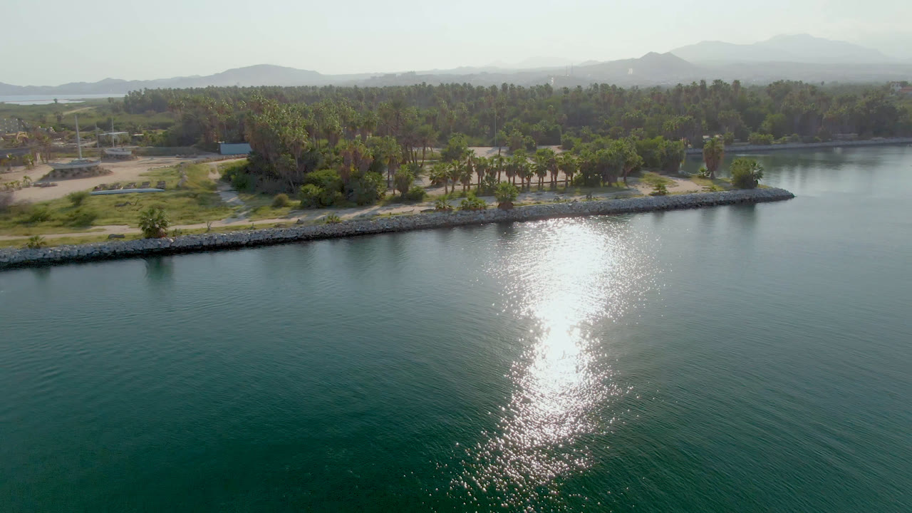 Aerial frame from Puerto Los Cabos showing green-blue water, a vertical sun reflection, a rock-lined shore, palms, low buildings, hazy hills, and no visible people.