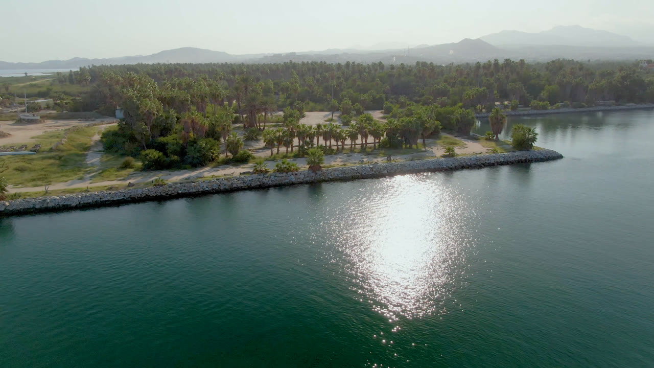 Aerial shoreline view with calm water, palms, a small lighthouse, and hazy mountains.