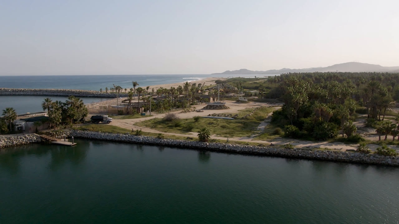 A wide aerial frame in Puerto Los Cabos shows dark lagoon water in front of a rock shoreline, palms, sandy ground, ocean, and hazy hills, with no visible people.