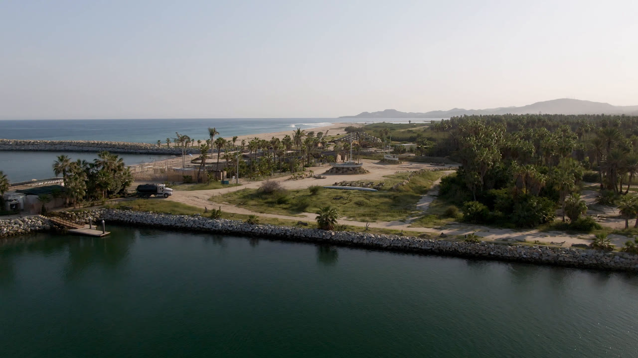 Opening frame of Puerto Los Cabos, Baja California Sur, with dark green water in front, a rock-lined shore, palms, beach, ocean, hazy hills, and no visible people.
