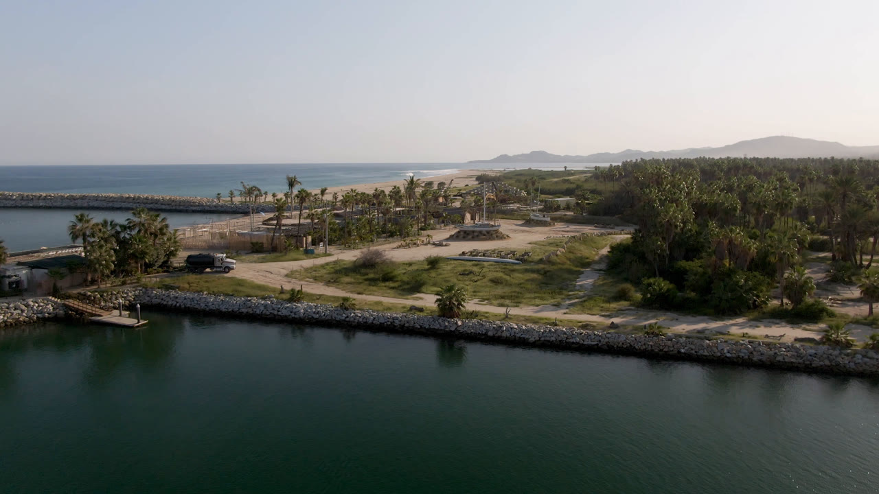 At 0:01 in Puerto Los Cabos, the drone-height view holds on calm water, a small dock at the lower left, palm clusters, sandy tracks, ocean, distant hills, and no visible people.