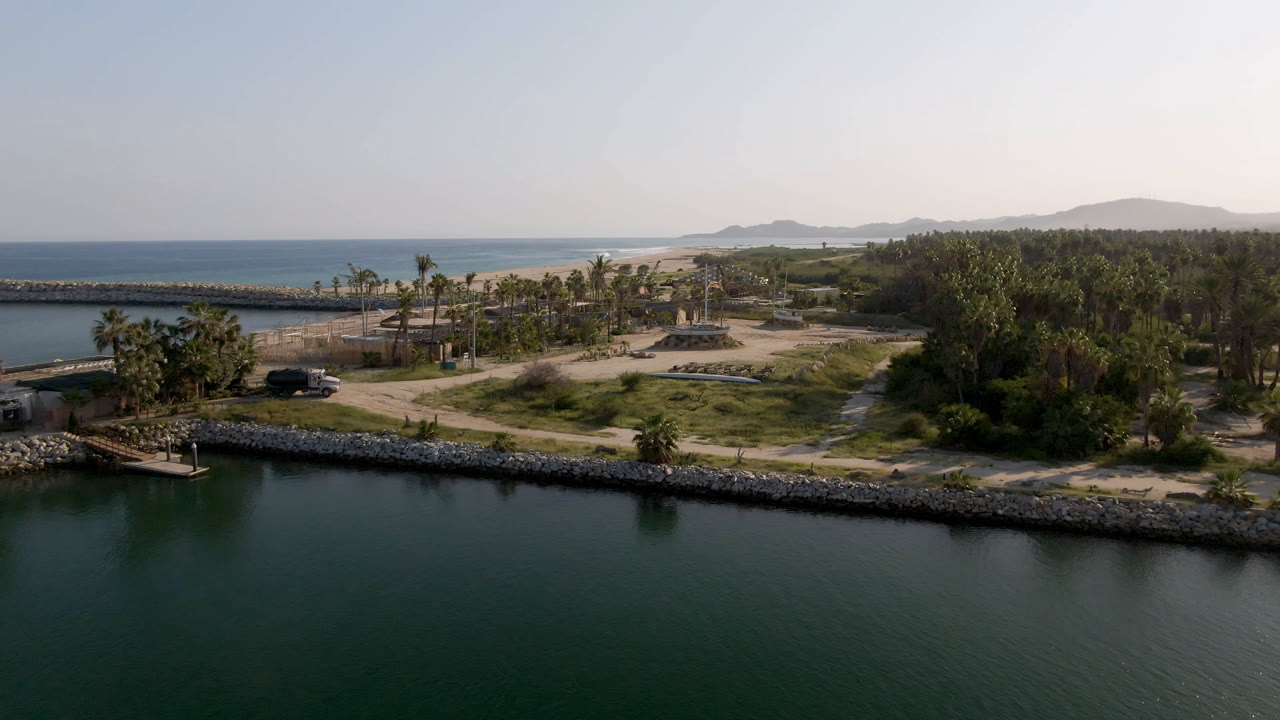 At 0:02, the Puerto Los Cabos frame keeps green water across the foreground, a rock seawall, palms and scrub on shore, pale beach and ocean behind, and no visible people.