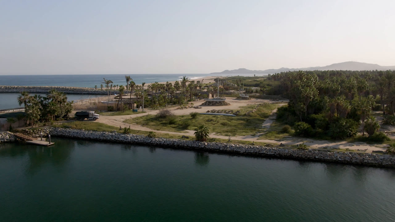 Aerial frame from Puerto Los Cabos showing dark inlet water, a rock shoreline, palms, dirt roads, blue ocean beyond the breakwater, hazy hills, and no visible people.