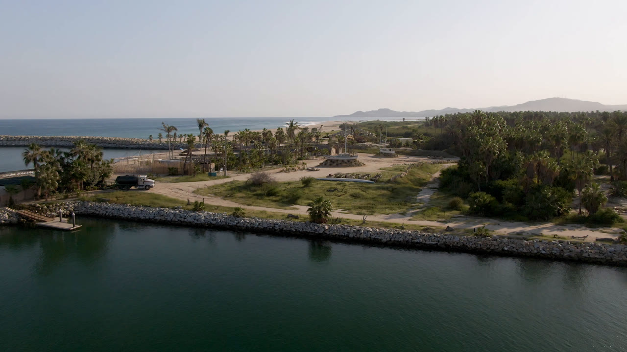 Wide aerial frame in Puerto Los Cabos with the inlet occupying the lower frame, a stone edge along shore, palm clusters near dirt roads, ocean in the distance, and no visible people.